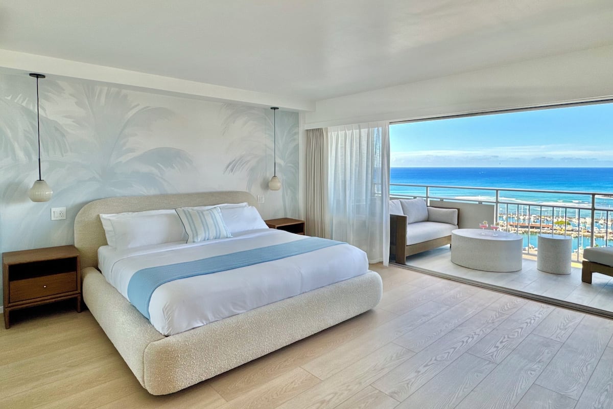A modern hotel room with a large bed, a balcony with a view of the ocean, and a seating area with a table.