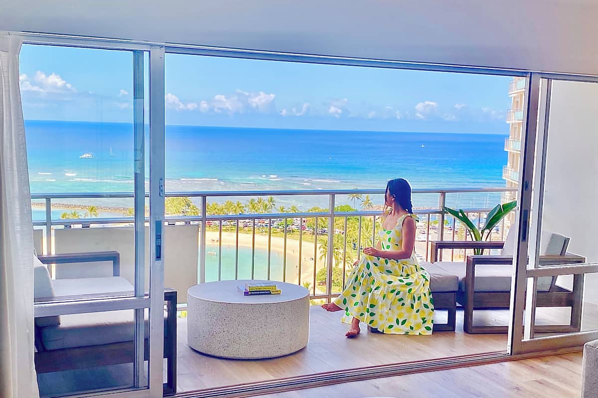 A balcony with a view of the ocean, a round table with a book, and a woman in a yellow dress.