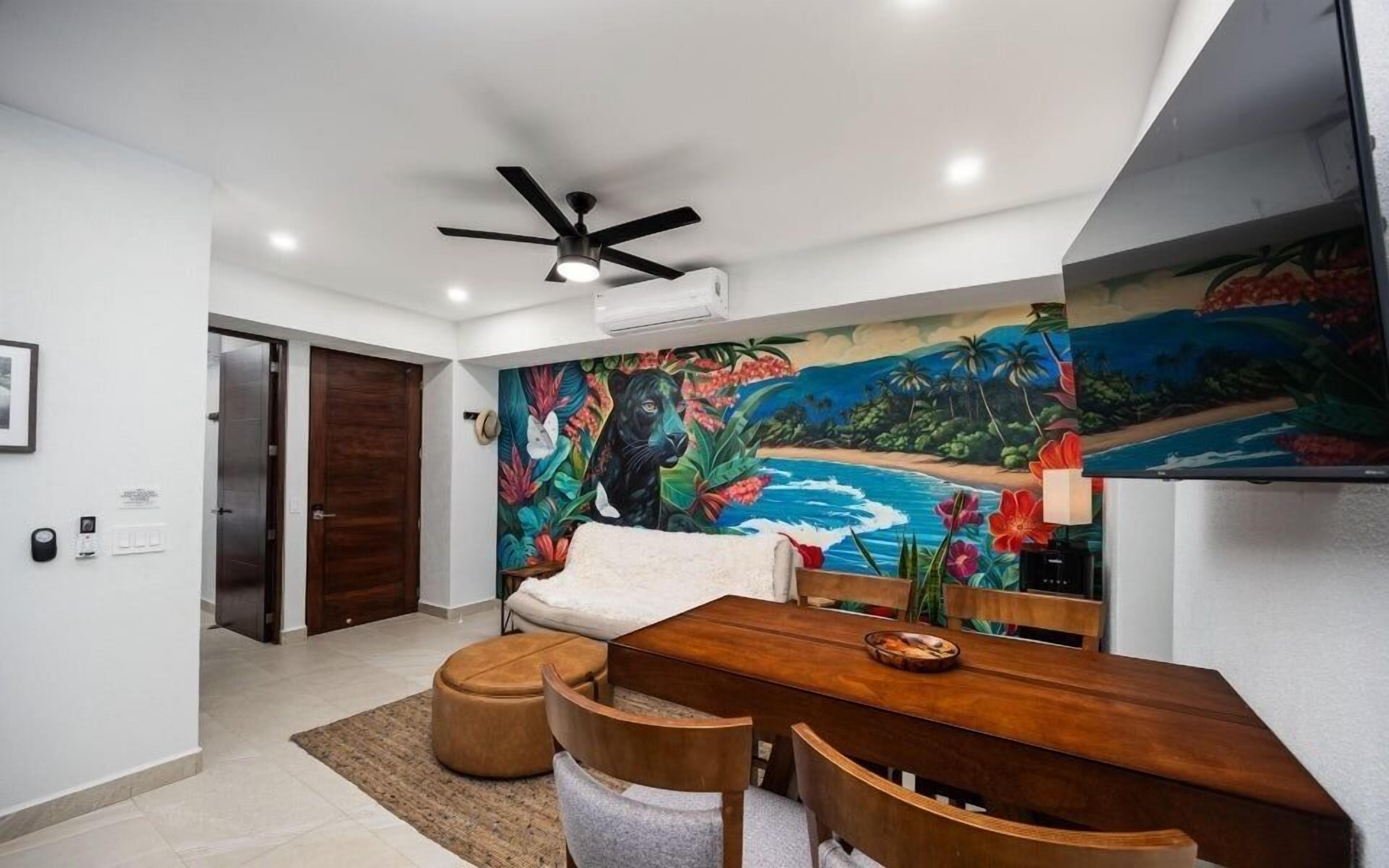 A dining area with a wooden table and chairs, a ceiling fan, and a vibrant tropical mural on the wall.