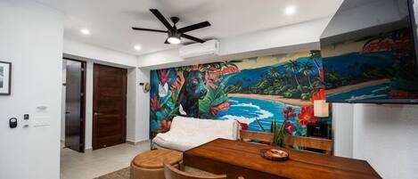 A dining area with a wooden table and chairs, a ceiling fan, and a vibrant tropical mural on the wall.