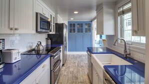 A modern kitchen with white cabinetry, blue accents, stainless steel appliances, and a large window providing natural light.