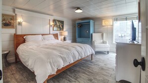 A bedroom with a wooden bed, white walls, a blue cabinet, a white chair, and a window with blinds.