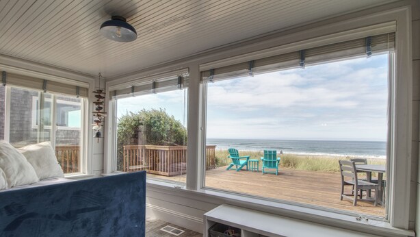 A room with a large window overlooking a beach, a blue sofa, and a wooden deck with outdoor furniture.