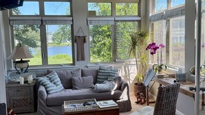 A spacious living room with a grey sofa, a wooden coffee table, and a wicker chair. Large windows provide ample natural light and views of greenery.