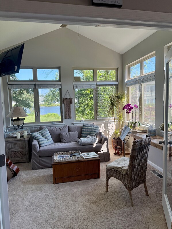 A spacious living room with a grey sofa, a wooden coffee table, and a wicker chair. Large windows provide ample natural light and views of greenery.
