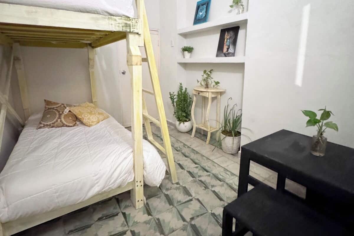 A small, compact room with a bunk bed, a small table, and a shelf with plants and pictures.