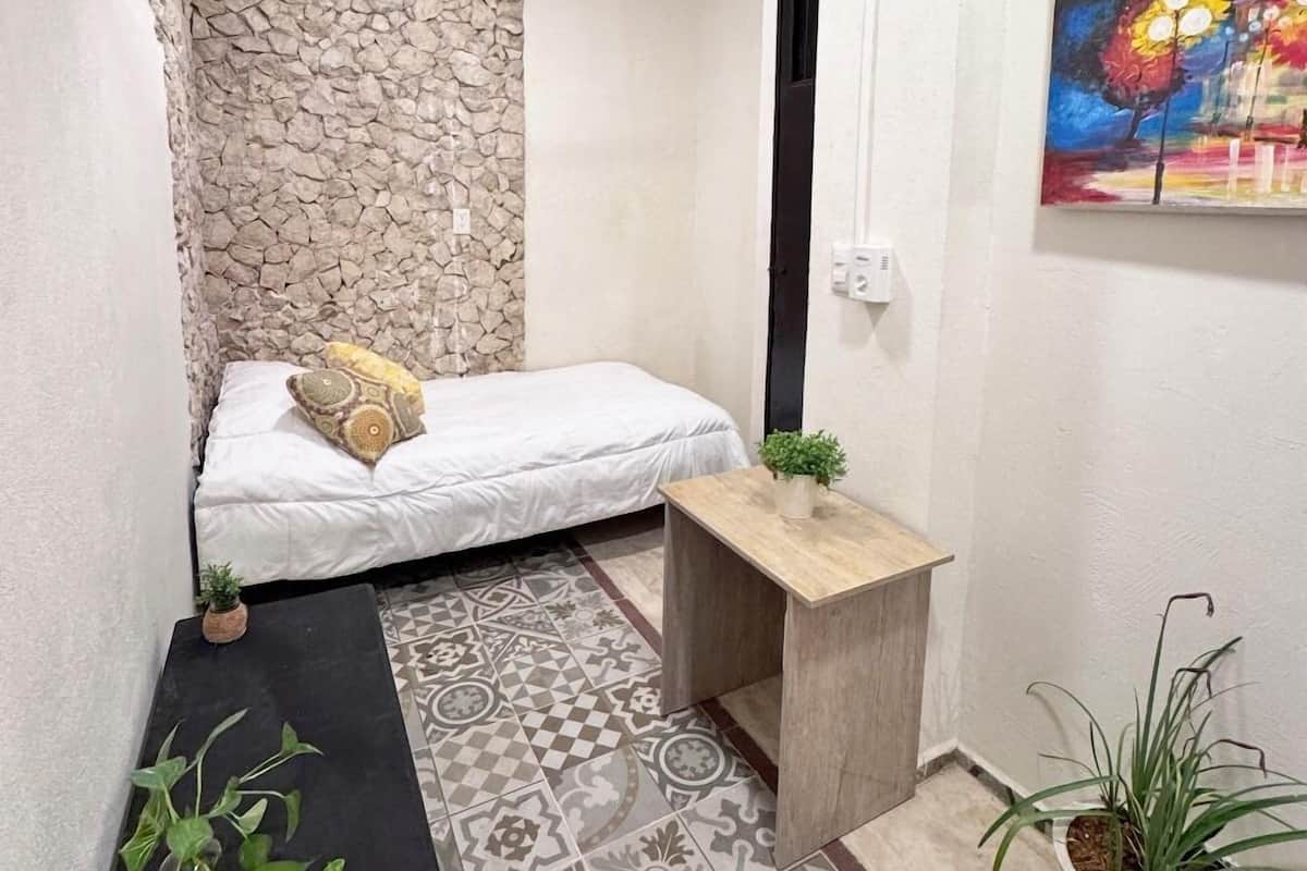 A small, narrow room with a bed, a small table, and a potted plant.