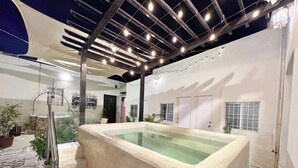 Et spa-område med et stort, rektangulært spabad, en pergola med lyskæder og en stenvæg.