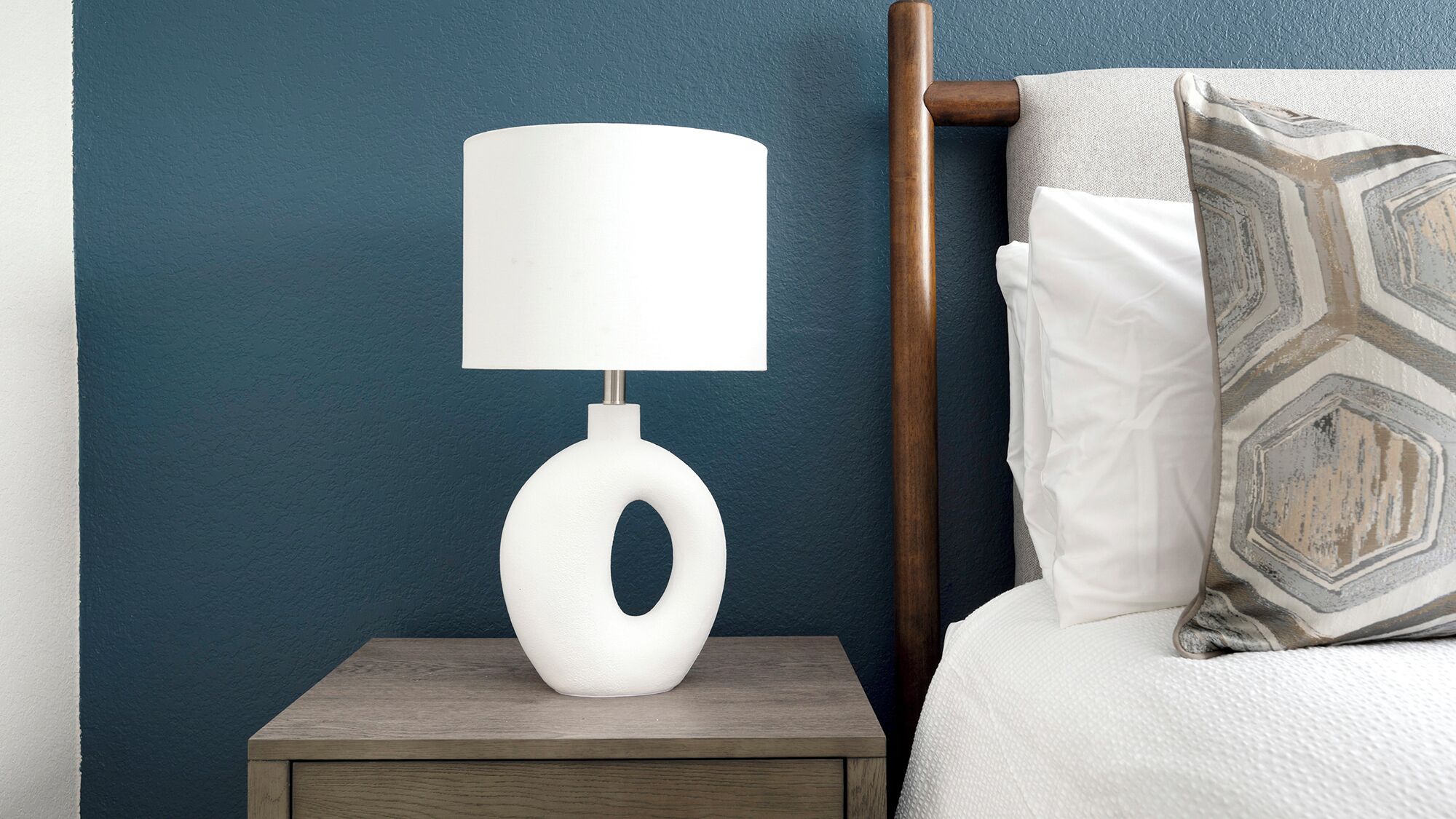 A bedside table with a white lamp and a patterned pillow on a bed against a blue wall.