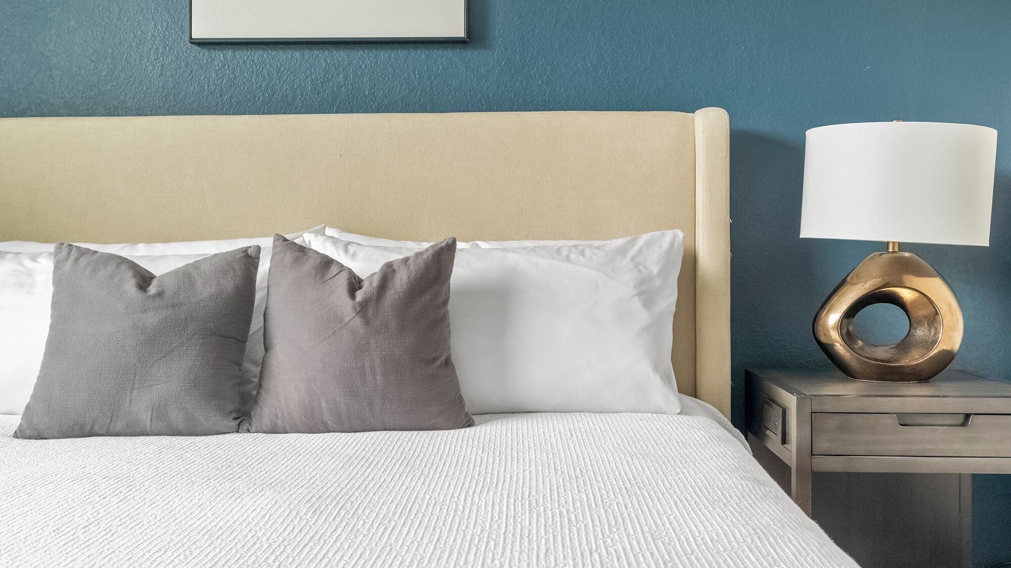 Un lit avec une literie blanche et deux oreillers gris, une tête de lit beige, une table de chevet avec une lampe moderne et un mur orné d’un tableau encadré.