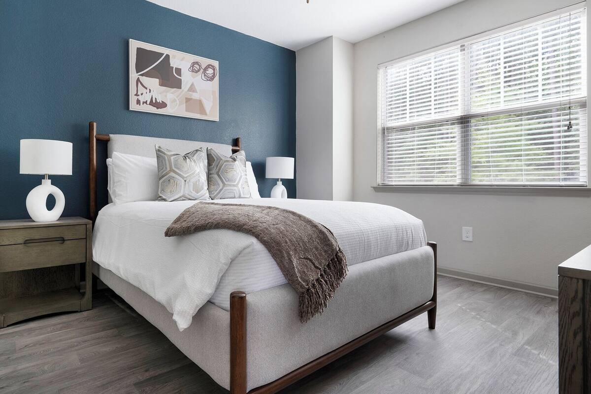 A bedroom with a bed, nightstands, a window with blinds, and a wall with a painting.