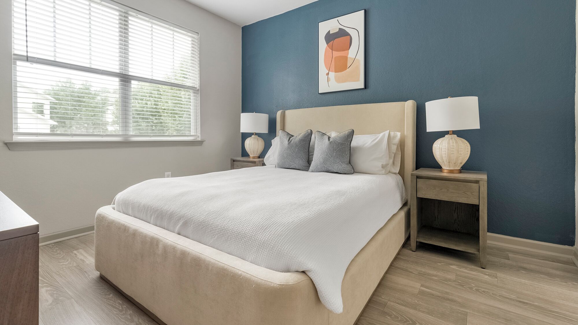 A neatly arranged bedroom with a large bed, two bedside tables with lamps, a wall-mounted artwork, and a window with blinds.