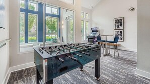 Een moderne kamer met een tafelvoetbaltafel, grote ramen en een zithoek met stoelen en een tafel.
