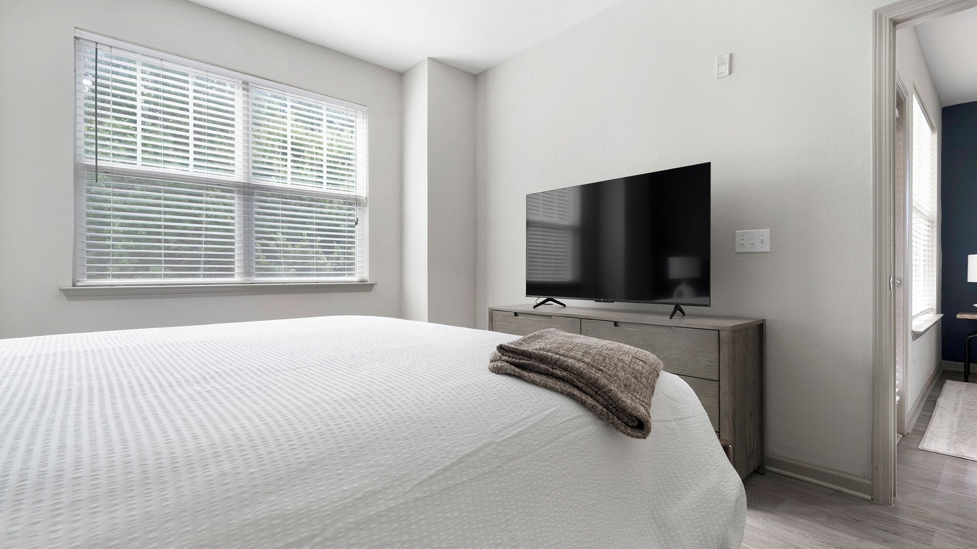 Une chambre à coucher avec un lit, un téléviseur posé sur une commode et une fenêtre avec des stores.