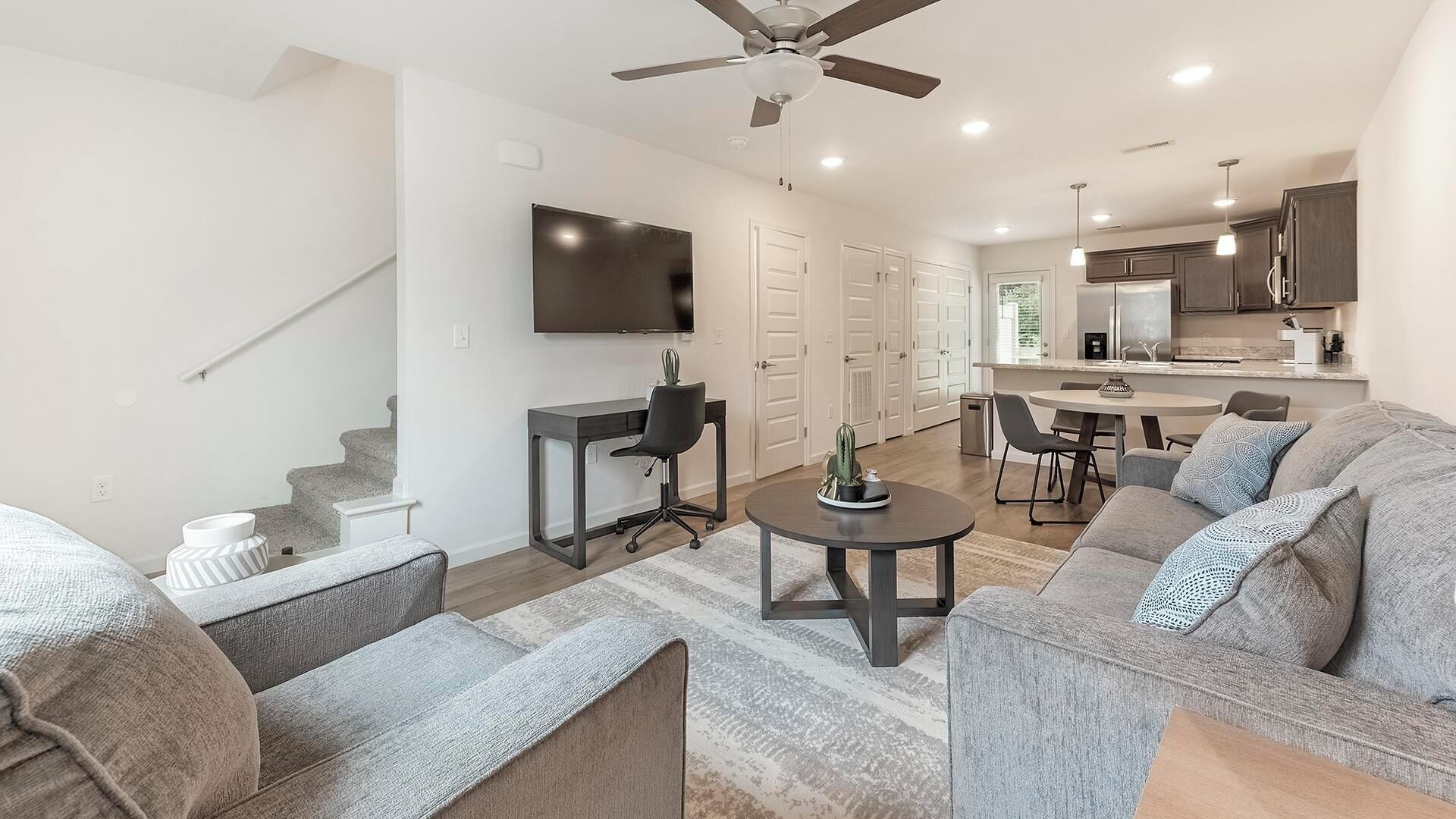 A modern living room with a gray sofa, a coffee table, and a TV. There is a staircase leading to another level, a kitchen area with a dining table, and a ceiling fan.
