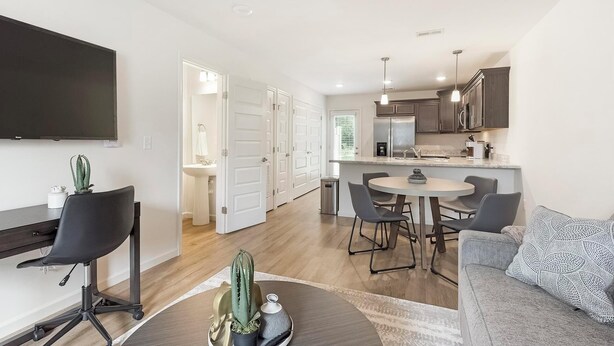 A modern living room with a flat-screen TV, a dining area with a round table and chairs, a kitchen with dark cabinets and a refrigerator, and a sofa with a patterned pillow.