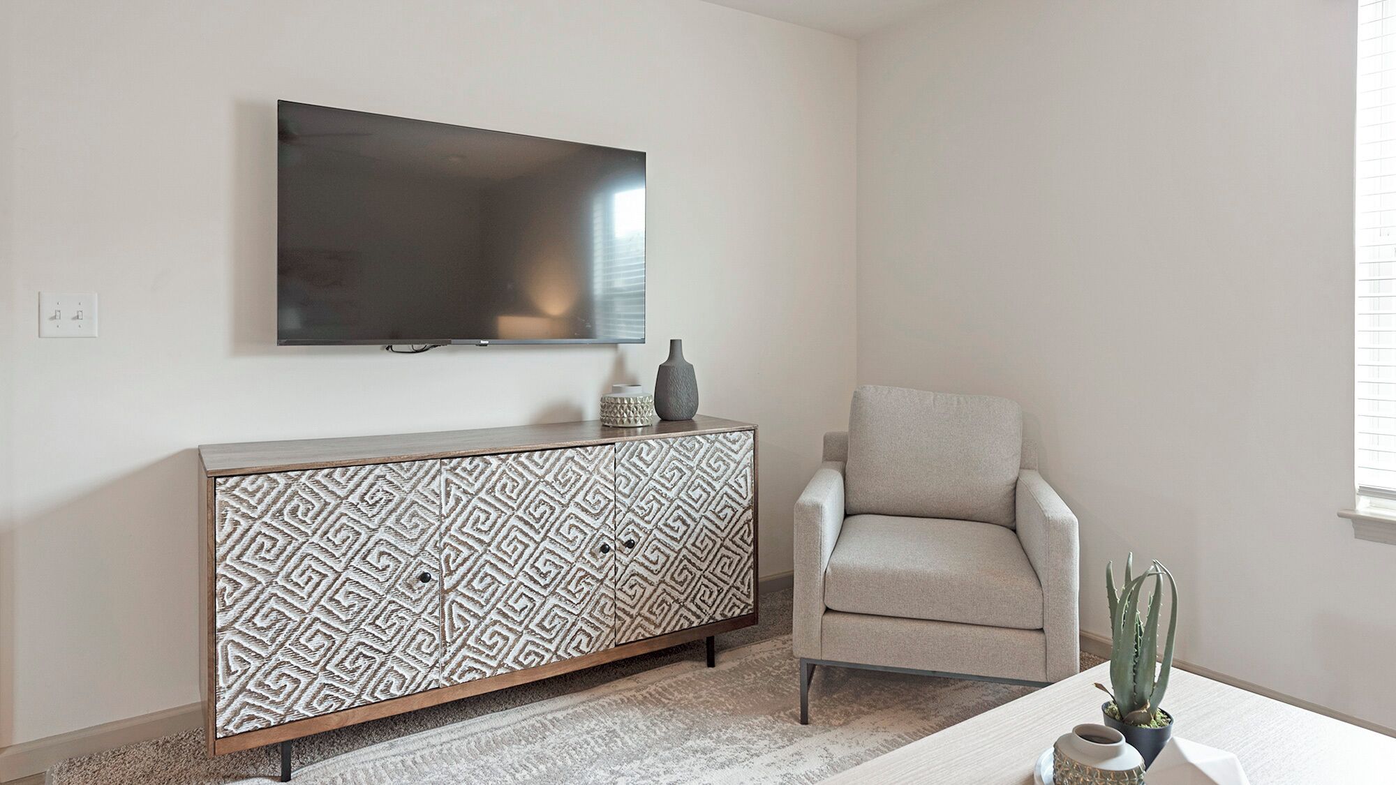 A living room with a flat-screen TV mounted on the wall, a patterned cabinet, a beige armchair, and a small table with a cactus plant.