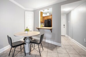 Un coin repas avec une table ronde et quatre chaises, une cuisine avec des meubles en bois et un réfrigérateur noir, et un couloir avec une porte blanche.
