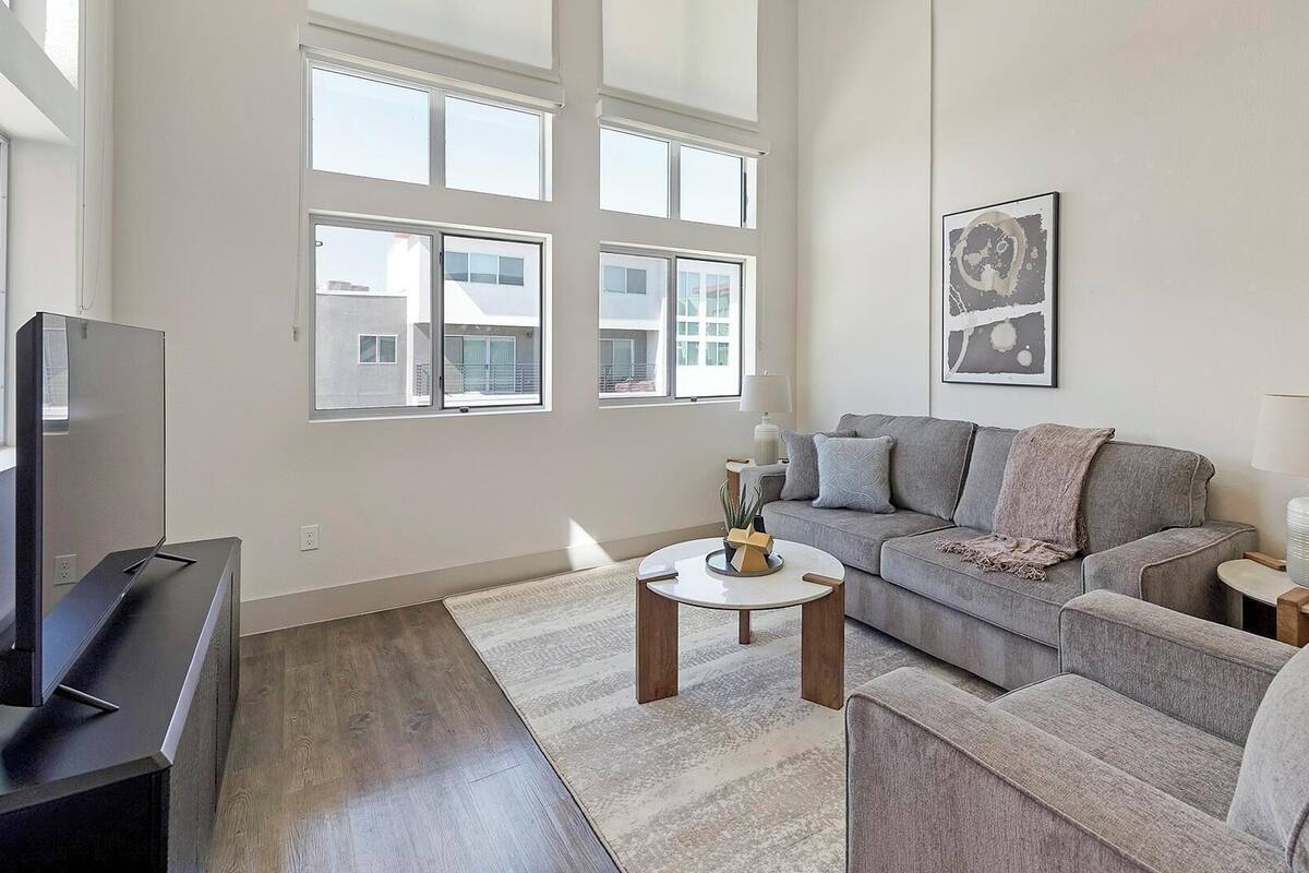 A spacious living room with a grey sofa, a coffee table, and a TV. Large windows allow natural light and offer views of buildings outside.