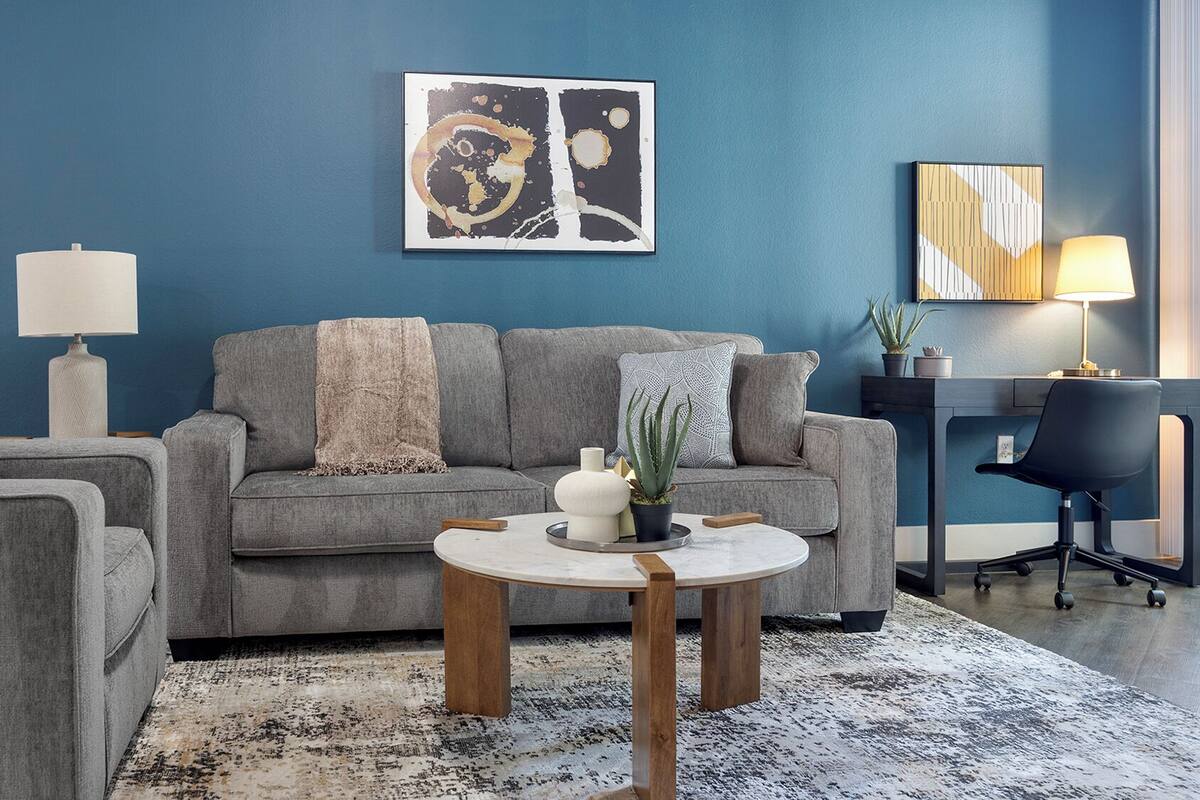 A modern living room with a blue accent wall, grey sofa, round coffee table, and a desk with a lamp.