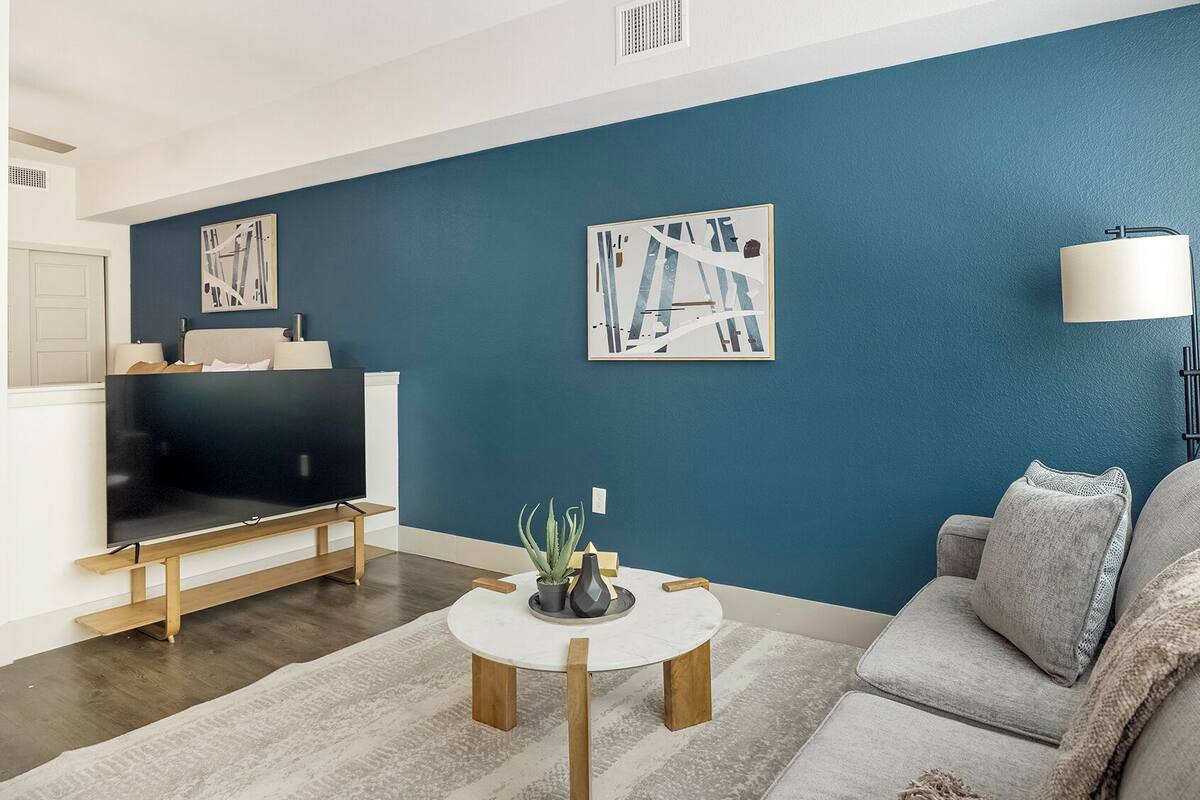 A living room with a dark blue wall, a flat-screen TV on a wooden stand, a round coffee table with a plant, and a grey sofa.