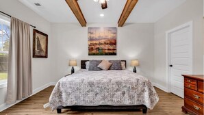 Une chambre à coucher comprenant un lit, deux tables de chevet, un ventilateur de plafond et une grande fenêtre avec des rideaux.