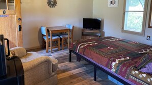 A room with a bed, a television, a wooden desk with a chair, a clock on the wall, and a window with blinds. - Stoney River Lodge - Unit 8 - Buffalo (Loveland)