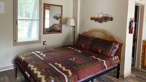 A bedroom with a wooden ceiling, a bed with a patterned quilt, a bedside lamp, a wall-mounted mirror, and a fire extinguisher. - Stoney River Lodge - Unit 8 - Buffalo (Loveland)