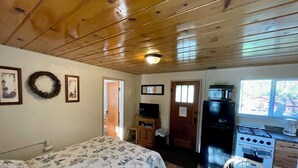 A cozy cabin with a wooden ceiling, a bed with a floral bedspread, a kitchenette with a stove, and a dining area with a table and chair.