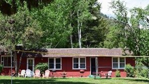 A red single-story house with a front porch, a lawn, and trees in the background.