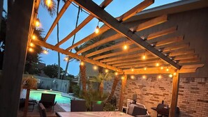 An outdoor patio with a wooden pergola, string lights, a pool, and a barbecue area.