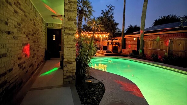 A swimming pool with green lighting, a brick wall, and palm trees.
