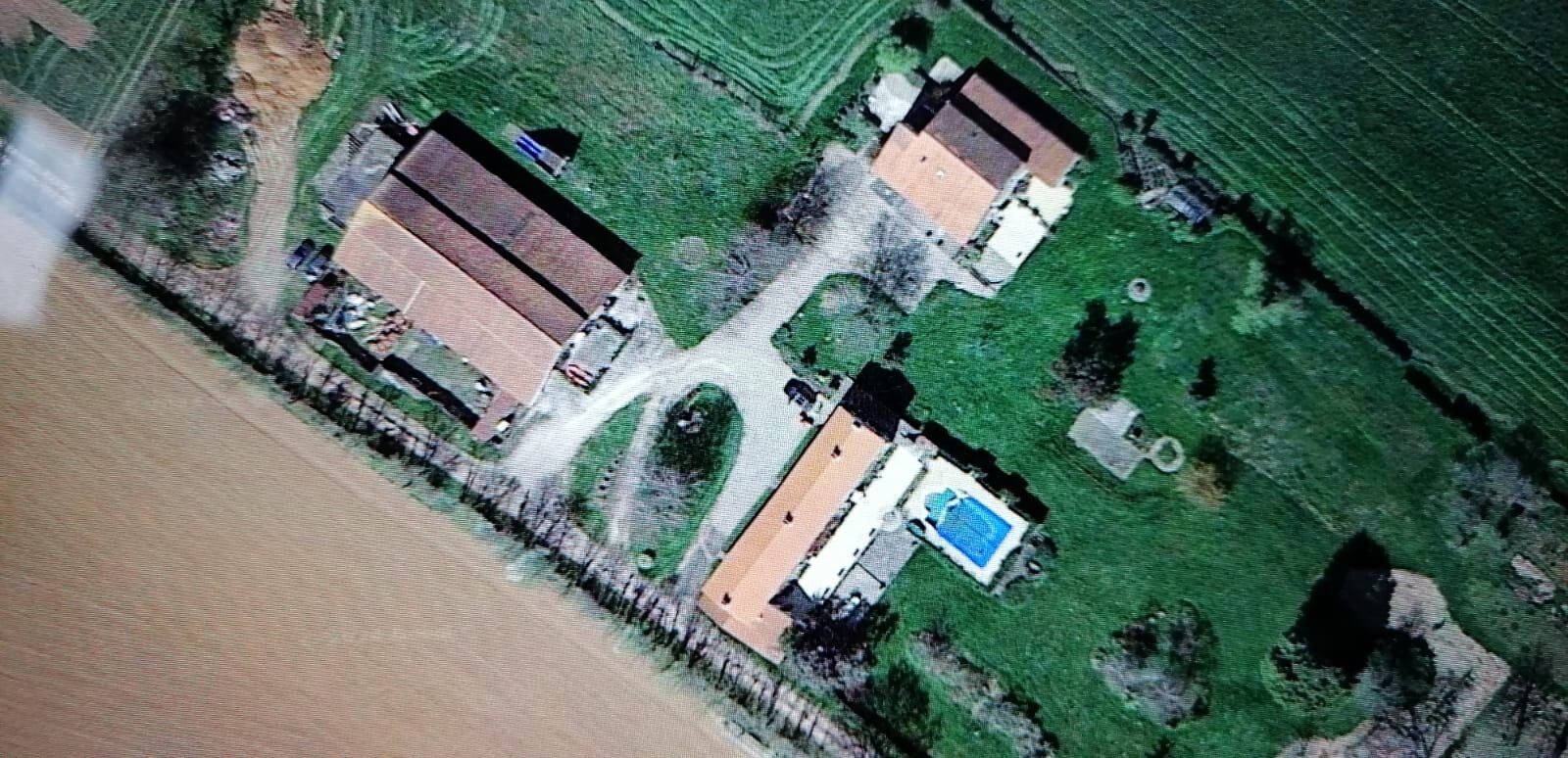 An aerial view of a property with two main buildings, a swimming pool, and surrounding green fields.