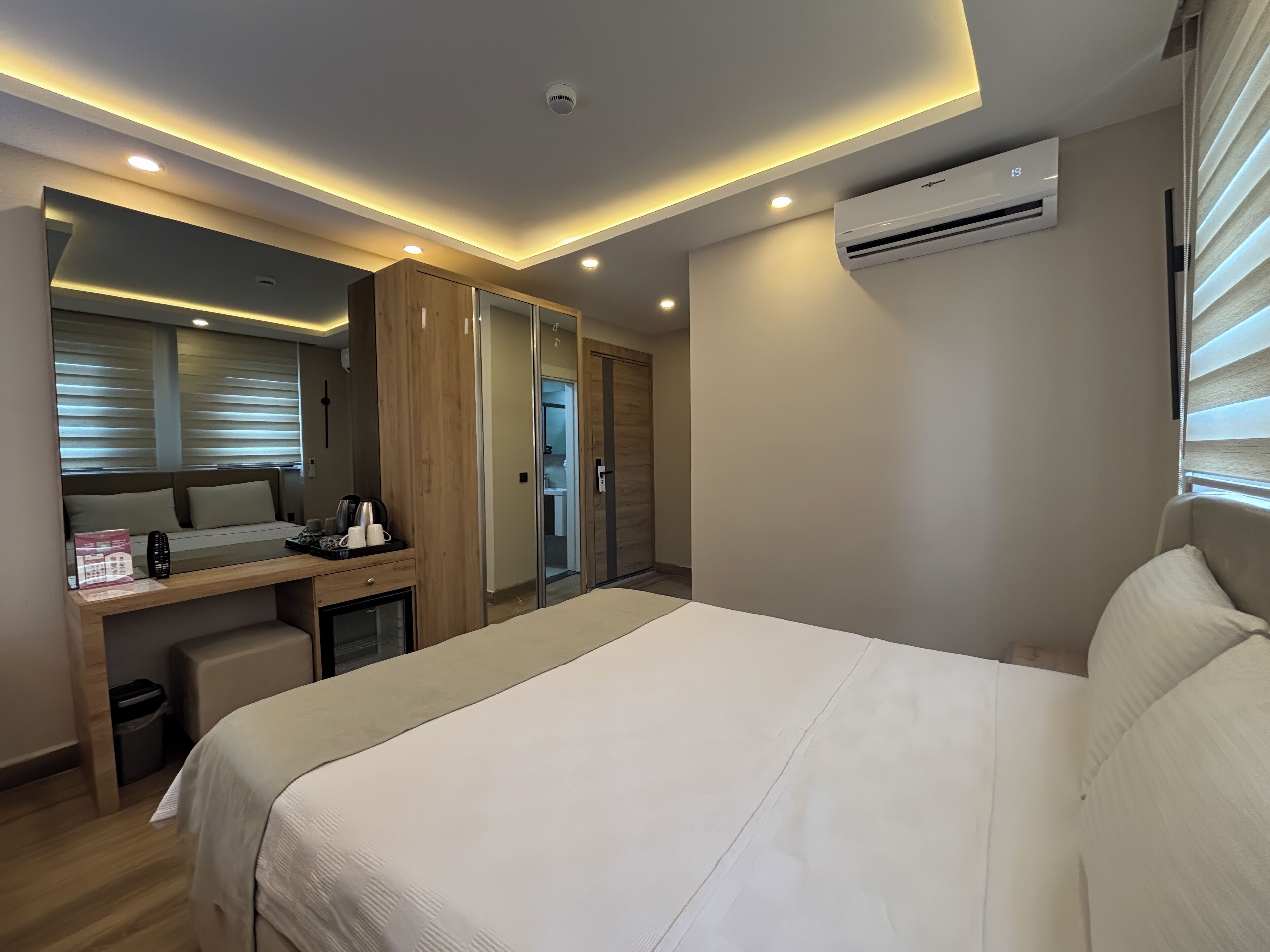 A modern hotel room with a large bed, a desk with a computer, a wardrobe, and an air conditioning unit.
