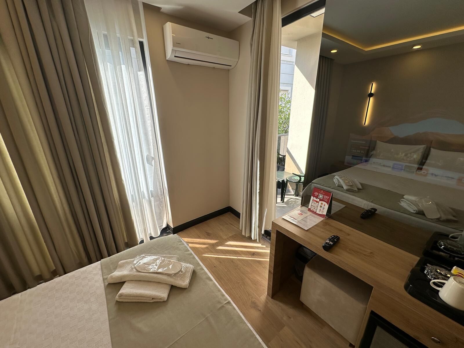 A hotel room with a bed, a desk with a telephone and remote control, a window with curtains, an air conditioning unit, and a view of the outside.