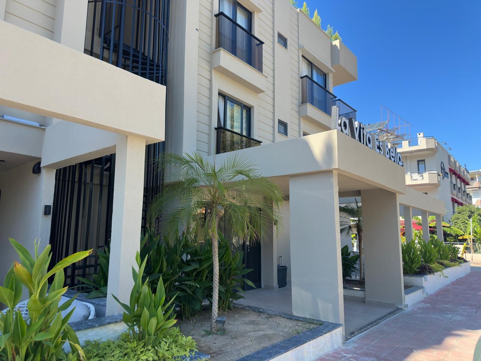 Un hotel moderno con un espacio exterior cubierto, palmeras y un cielo azul despejado.