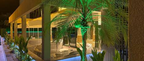 A well-lit outdoor area with a palm tree, lounge chairs, and a pool.