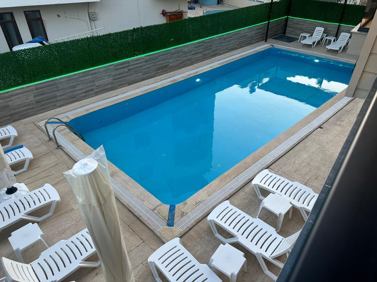 A rectangular pool with a tiled deck, surrounded by white lounge chairs and a green hedge.