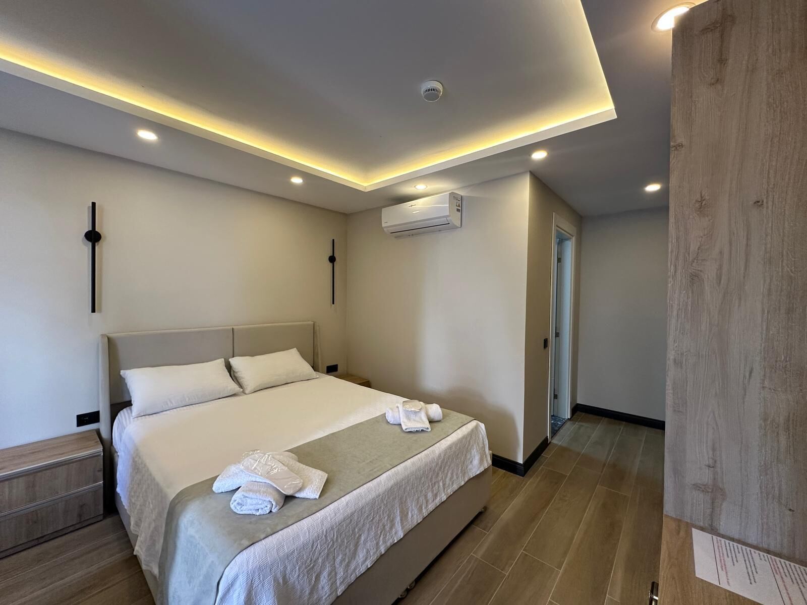 A modern hotel room with a large bed, a bedside table, an air conditioning unit, and a wooden panel on the wall.