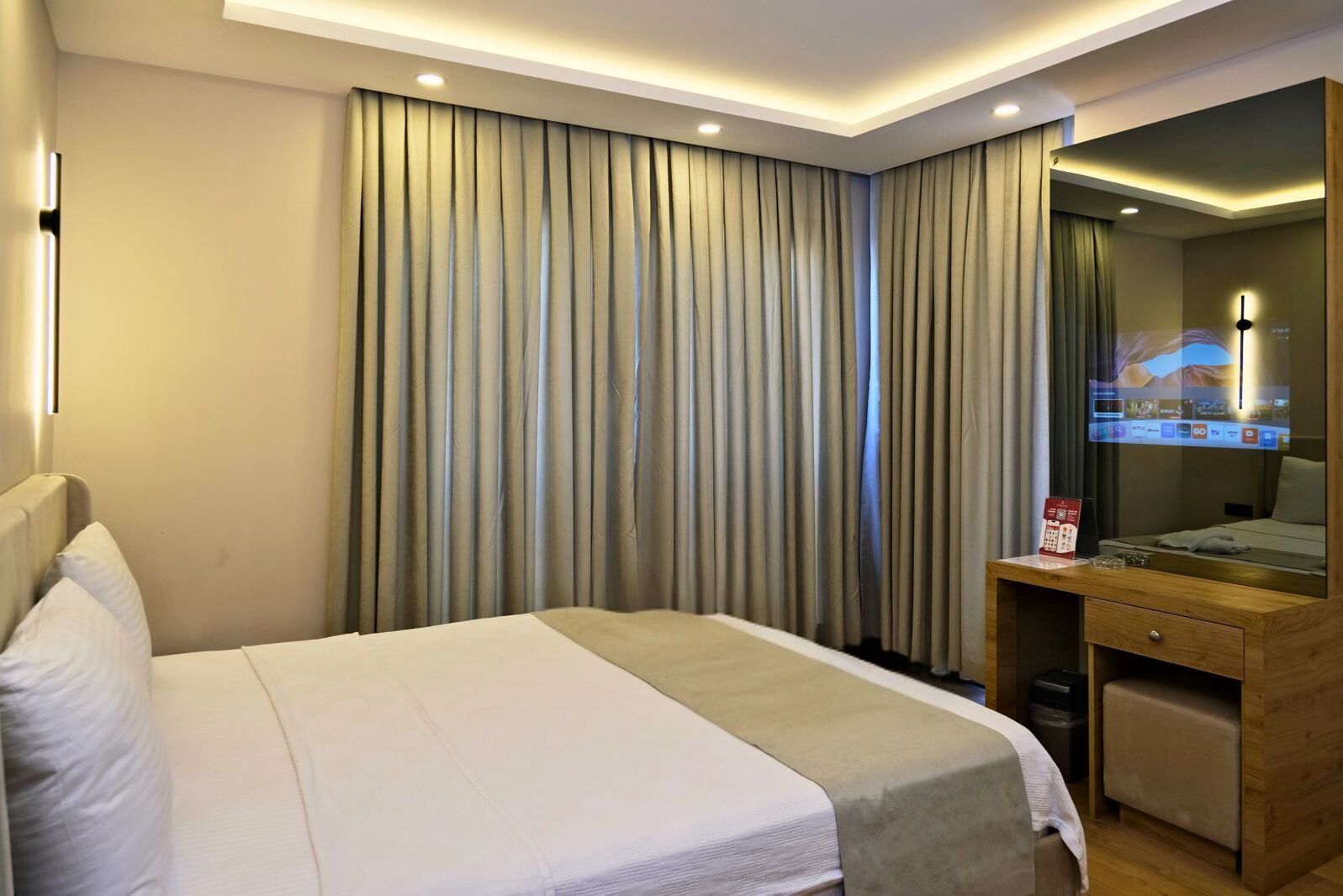Ein modernes Hotelzimmer mit einem großen Bett, einem hölzernen Schreibtisch mit Fernseher und einem großen Fenster mit Vorhängen.
