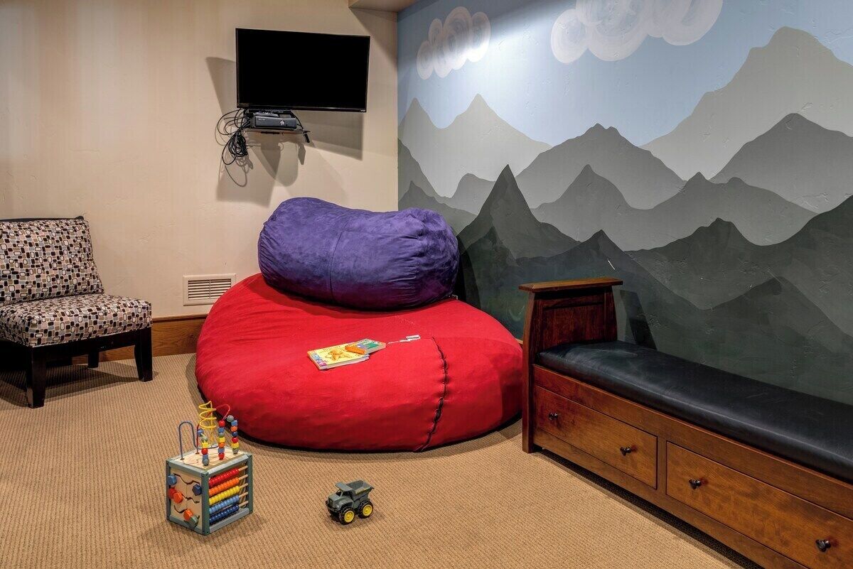 Una habitación con un mural de montañas, un sillón de peluche rojo, un armario de madera, un televisor montado en la pared y una silla estampada.