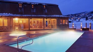 Ein Gebäude mit Poolanlage im Abenddämmerung, mit großem Pool, überdachter Sitzgelegenheit und Blick auf einen verschneiten Hügel.