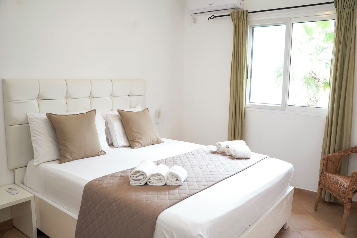 Una habitación de hotel con una cama grande, cabecera, ventana con cortinas, una silla y una mesita de noche.