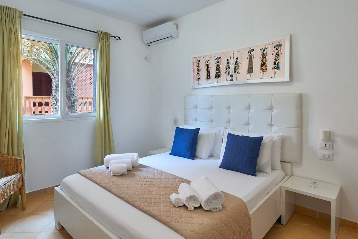 Habitación de hotel con cama, una silla, ventana con cortinas, aire acondicionado montado en la pared y un cuadro sobre la cama.