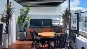 A rooftop terrace with a wooden table and chairs, a grill, and potted plants. - Get one (Joao Pessoa)