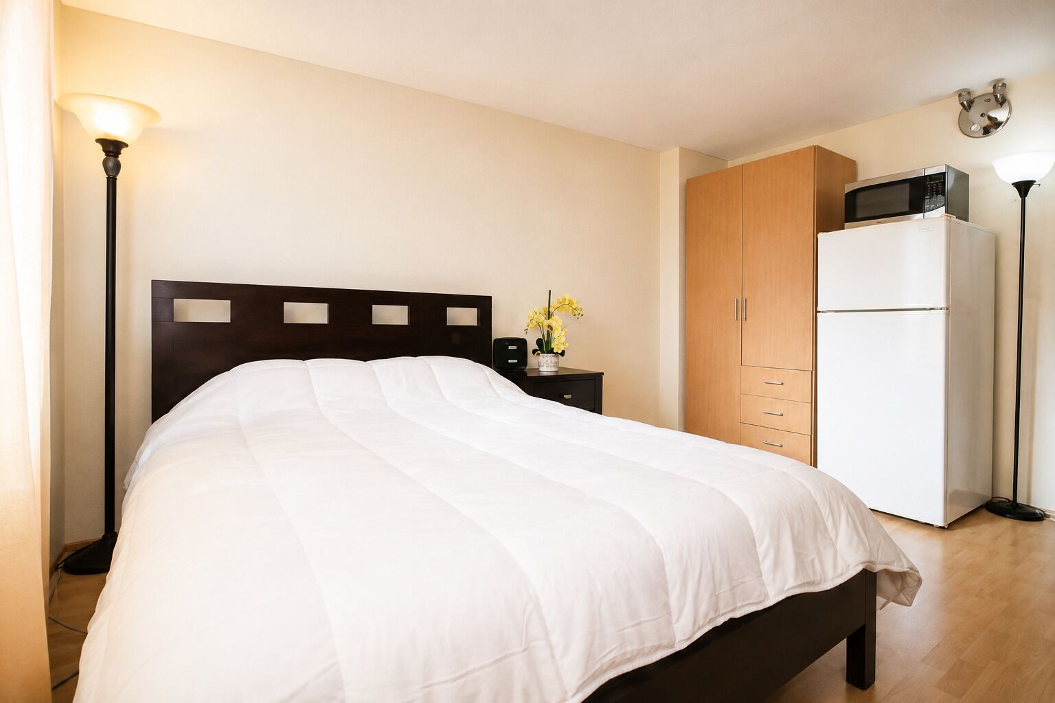 Un letto rifatto con cura in una camera d'albergo, con una testiera in legno, un comodino con una lampada, un piccolo vaso con fiori, un armadio e un frigorifero.