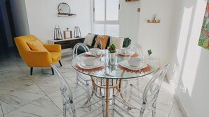 A modern dining area with a glass table and clear chairs, a yellow armchair, and a large window providing natural light. - The Point – Stylish 2-BR w/ Pool, Gym , Private Balcony & Free Parking (Panamá)