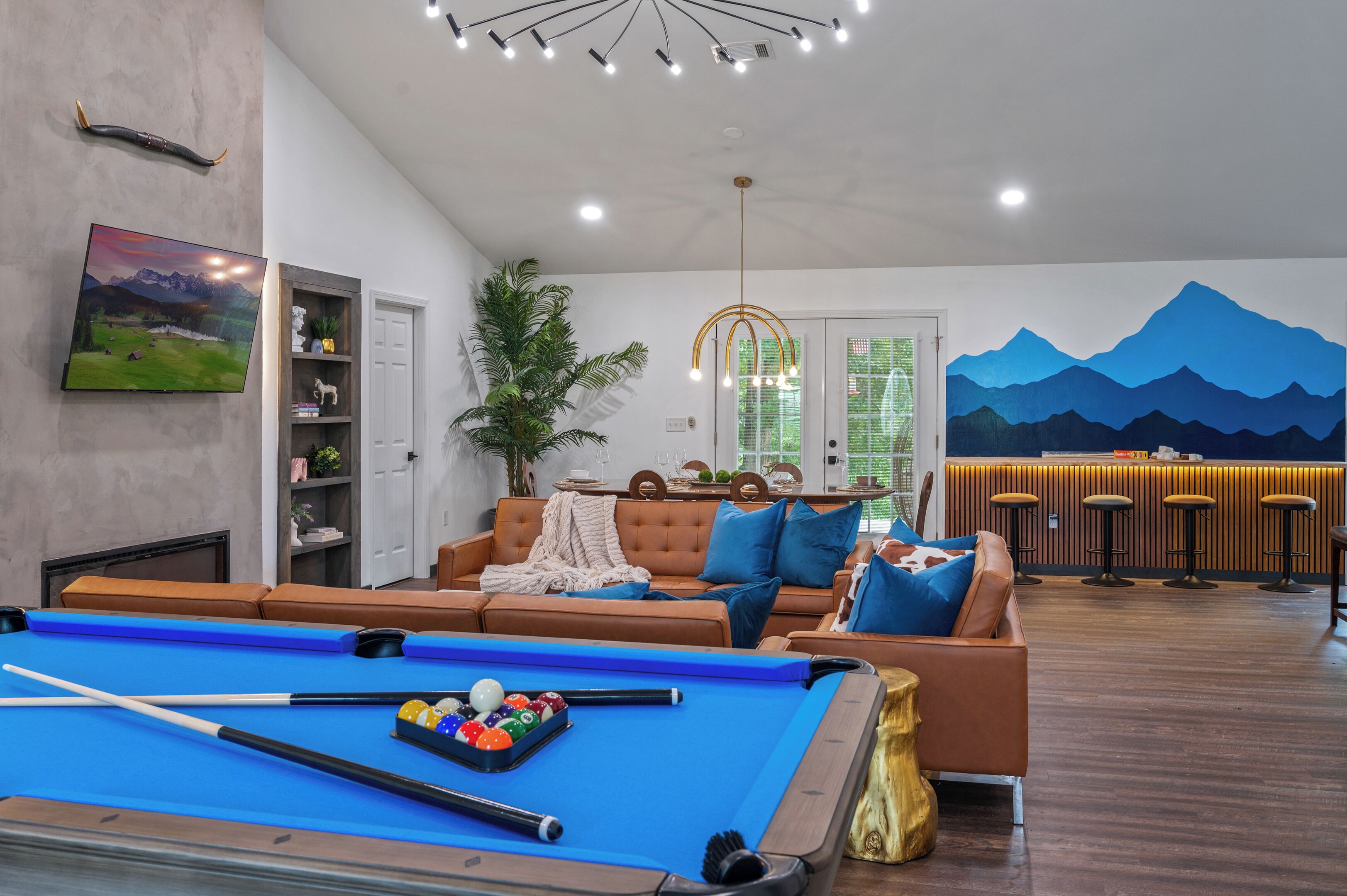 A modern living room with a pool table, a sofa, a bar area, and a mountain mural on the wall.