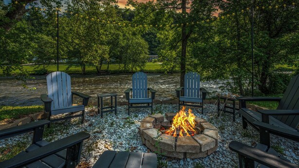 A fire pit surrounded by Adirondack chairs in a park setting with trees and a view of a house in the background. - River Front! Mini Golf! Hot Tub! Mountain views. (Sevierville)