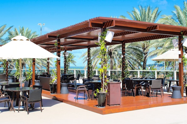 Eine Außenpatio mit Tischen und Stühlen, eine Holzpergola und Blick auf Palmen und den Ozean.